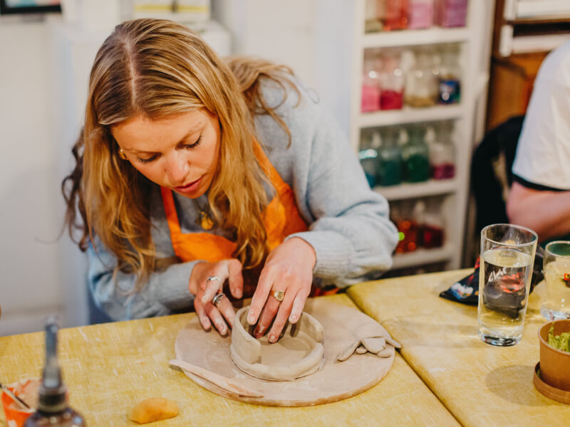 A woman wearing an orange apron works intently on some hand built pottery, touching the clay with her hands in a studio  